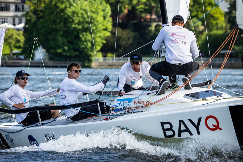 Pressemitteilung – Konstanzer Yacht Club 15. Mai 2023 Der Konstanzer Yacht Club segelt wieder in der 1. Segel-Bundesliga: Saisonstart in Berlin vom 12.-14. Mai 2023 - mit mutigem Nachwuchsteam Der Verein Seglerhaus am Wannsee war Gastgeber für den ersten Spieltag der Segel-Bundesligasaison 2023. Insgesamt 36 Teams in zwei Ligen kämpfen in dieser Saison an sechs bzw. fünf Spieltagen um die begehrte Meisterschale, SAILING Champions League Qualifikation, den Klassenerhalt oder den Aufstieg. Finale ist im Oktober in Hamburg auf der Außenalster. Der erste Spieltag der neuen Saison 2023 fand vom 12. bis 14. Mai auf dem Wannsee statt. Für je 18 Vereine in der 1. und 2. Segel-Bundesliga ging es wieder um Punkte, Qualifikationen und für den einen oder anderen auch um den Abstiegskampf. Nicht so für den KYC: Nach einer spannenden Saison 2022 gelang den Teams des Konstanzer Yacht Club der stabile Aufstieg von der zweiten in die erste Bundesliga. Noch eine Woche zuvor wurde dieser von den aktiven Seglern, Sponsoren und Vereinsmitgliedern mit einem leckeren Grillbuffet der neuen Wirte, Lilli Reifschneider und Axel Seitz, auf der Terrasse des Clubhauses gefeiert. Bereits sechs Tage später brach die erste Crew Richtung Berlin auf, um sich auf bundesdeutschem Niveau zu messen und präsentieren. Steuermann Bendix Mayer, Nicolas Kutter als Taktiker, Nils Hoffmann, zuständig für den Trimm, und Jakob Zürn auf dem Vorschiff starteten als neu formiertes Team zu einer ersten sportlichen Standortbestimmung, die in der Segelwelt aufmerksam verfolgt wird. Gemeinsam haben die vier ein Durchschnittsalter von knapp 20 Jahren und stellen ein ehrgeiziges und hoch motiviertes Nachwuchsteam dar. „Selten waren die Teams vor dem ersten Spieltag so gut vorbereitet und trainiert“, so heißt es in der Pressemitteilung der Segelbundesliga. Das gilt auch für das Konstanzer Team. Die Trainingsphase begann bereits zu Ostern, als sich mehrere Liga-Teams am Gardasee trafen und mit einem Top-Trainer ihre Manöver optimierten. Doch trotz aller Vorbereitungen ist der Wannsee mit seinen speziellen Windverhältnissen immer wieder eine Herausforderung. Nach einem klaren ersten Platz im ersten Rennen war das Team einige Wettfahrten lang vom Pech begleitet. Es folgte eine Serie von letzten und vorletzten Zieleinläufen, für die es eigentlich keine eindeutige Erklärung gibt. Weder Frühstarts, noch Vorfahrtsprobleme oder technische Fehler unterliefen dem Team. Beachtenswert, dass es der Crew gelang sich nach dieser Phase wieder stabil in das Mittelfeld zurück zu kämpfen und sogar noch zwei weitere erste Plätze zu segeln. Das erfordert eine mutige Taktik, stabiles Selbstvertrauen und auch ein wenig Frechheit im richtigen Moment. So wurde der Saisonstart in der Bundesliga mit Rang 14 beendet, eine gute Ausgangsbasis für die weiteren Events. Die spannenden Rennen wurden wieder über das SAP-Tracking-System live abgebildet, teilweise sogar live gestreamt und konnten so von den Daheimgebliebenen gebannt verfolgt werden. Bis zum Saisonende stehen noch fünf weitere Regattawochenenden an, bei denen der KYC sich in der ersten Bundesliga behaupten kann. Zweimal finden die Events sogar am Bodensee statt: vom 15.-17. September direkt in Konstanz, mit dem KYC als ausrichtenden Verein, und dann noch einmal im Oktober in Überlingen. (Karin Franke, Konstanzer Yacht Club) Die Crew des Konstanzer Yacht Clubs (Foto: Adam Burdyło) Das KYC-Team: Nils Hoffmann, Bendix Mayer, Jakob Zürn, Nicolas Kutter – von links (Foto: Adam Burdyło)